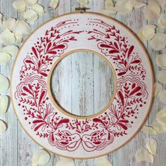 a red and white embroideryed wreath on a wooden surface with petals around it, ready to be framed