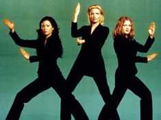 three women in black outfits are dancing against a green background with their hands up to the side