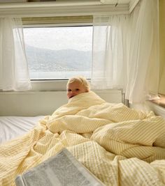 a little boy laying in bed under a blanket and looking out the window with an open book