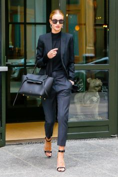 a woman in black suit and heels walking down the street with her hand on her hip