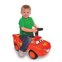 a young boy riding on the back of a red toy car with a green shirt