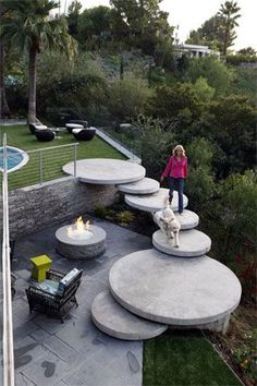 a woman standing on top of a cement bench next to a fire pit in the middle of a lush green park