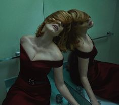 two women in red dresses are sitting on a bathroom sink and one is looking into the mirror