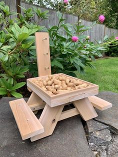 a wooden picnic table with peanuts in it on top of some rocks and flowers behind it