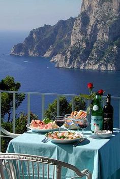 a table with food and wine on it overlooking the ocean in front of a cliff