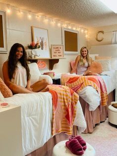 two women sitting on twin beds in a dorm room with lights strung above the bed