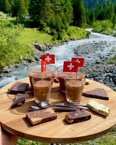 two glasses of chocolate with swiss flags on the table next to a river and mountains