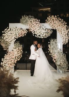 a bride and groom pose for a photo in front of a floral arch at night