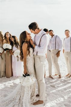 a bride and groom kissing in front of their wedding party on the beach at sunset