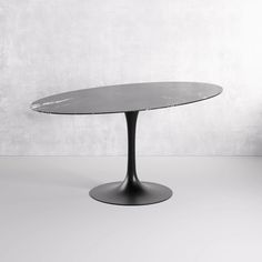 an oval glass table with black base on a white floor in front of a concrete wall