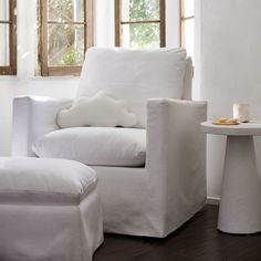 a white chair and foot stool in a room with two windows, one has a pillow on it