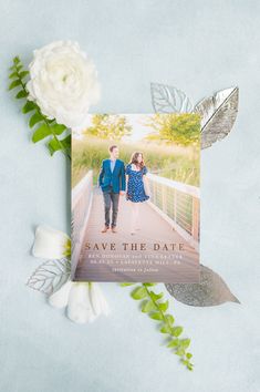 save the date card with white flowers and greenery on blue paper next to it