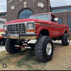 an old red truck parked in front of a building with large tires on it's flatbed