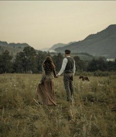 a man and woman holding hands walking through a field with mountains in the back ground