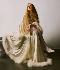 a woman with long blonde hair sitting on a white couch wearing a fur coat and high heels