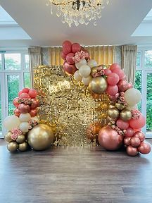 a large balloon arch with gold, pink and white balloons on the floor in front of a chandelier