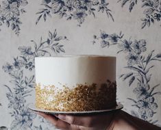 a person holding a cake with gold sprinkles on it in front of a floral wallpaper