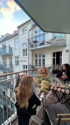 three women sitting at an outdoor table on a balcony overlooking the city with buildings and balconies