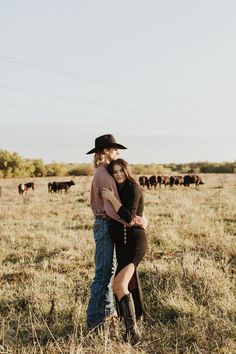 a man and woman standing in the middle of a field with cows behind them on a sunny day