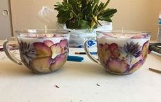 two cups with flowers painted on them sitting on a table next to a potted plant