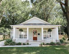 a small white house with red door and porch in the middle of a tree - lined yard