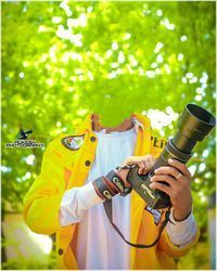 a man in yellow jacket holding a camera and looking into the distance with trees behind him