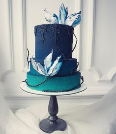 a three tiered cake with blue frosting and flowers on top, sitting on a pedestal