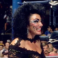 a woman with black hair and face paint smiles at the camera in front of an audience