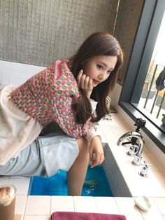 a woman sitting on the edge of a bathtub in front of a window next to a sink