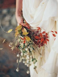 a woman in a white dress holding a bouquet of leaves and flowers with her hands