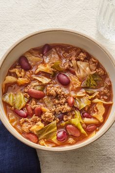 a bowl of soup with meat, beans and vegetables on a white cloth next to a glass of water