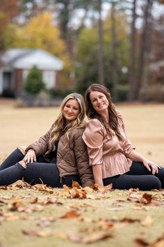 two beautiful women sitting on the ground in front of some trees and leaves with their arms around each other