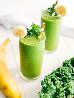 two glasses filled with green smoothie next to bananas and broccoli