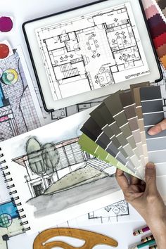 a person's hand holding color samples in front of architectural drawings and design tools