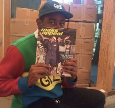 a man sitting on the ground holding up a magazine in front of his face and wearing a hat