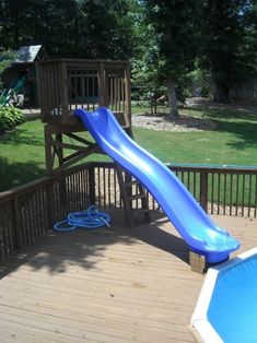 a blue slide in the middle of a wooden deck next to a swimming pool and tree