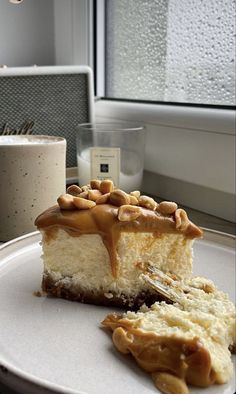 a piece of cheesecake on a white plate next to a cup and window sill