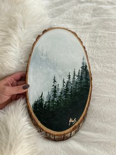 a person holding up a painting on top of a wooden slice with trees in the background