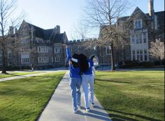 two people walking down a sidewalk in front of a large building with trees and grass