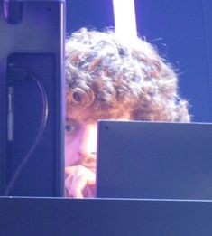 a young man with curly hair is looking out from behind a computer screen at the same time