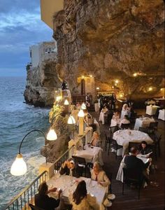 people sitting at tables in front of the ocean and cliff side restaurant with lights on