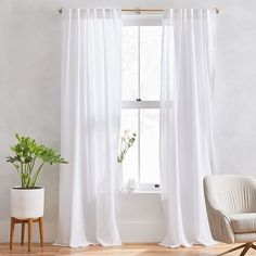 a living room with white curtains and a chair in front of the window, next to a potted plant