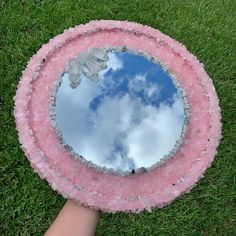 a person's hand holding a pink and white mirror with clouds in the sky