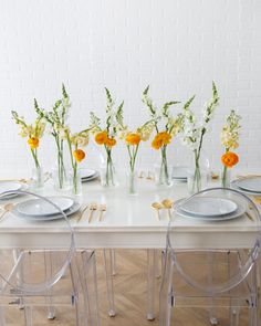 the table is set with clear chairs, plates and vases filled with yellow flowers