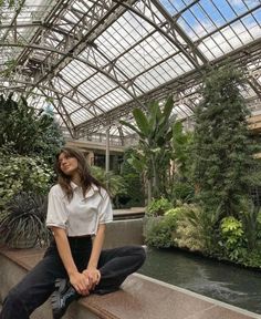 a woman sitting on the edge of a walkway next to a pond in a greenhouse