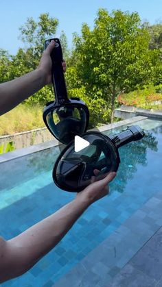 a person holding a camera in front of a swimming pool with trees and bushes behind it