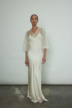 a woman standing in front of a white wall wearing a long dress with sheer sleeves