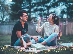 a man and woman sitting on a blanket blowing bubbles in the air while they sit next to each other