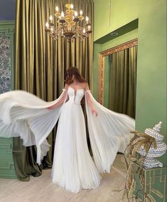 a woman in a white dress is standing near a chandelier with her arms outstretched