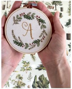 two hands holding up a cross stitch monogrammed wreath with the letter a on it